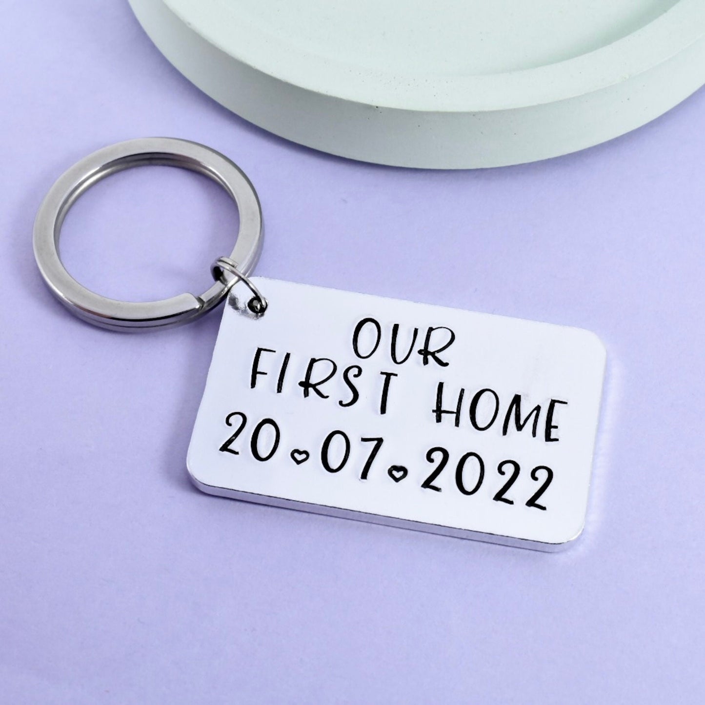 A silver keyring with a rectangular tag that reads 'OUR FIRST HOME 20.07.2022' on a purple background.