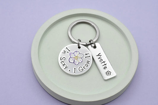 A personalised disc keyring with the text "I'm Sexy and I Grow It" with large purple flower design in the center. Also has a silver rectangle tag, personalised with a name and small black flower design