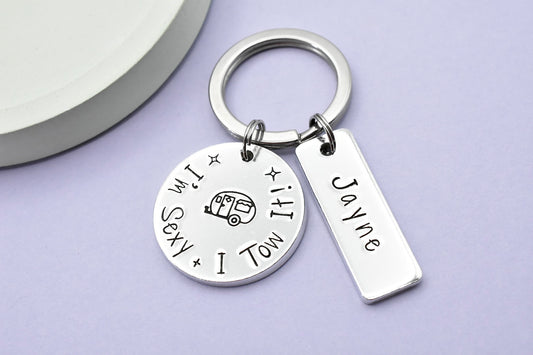 A silver keyring with a circular charm featuring the text 'I'M SEXY AND I TOW IT', and a rectangular tag with the name 'Jayne'. It is presented on a purple background.