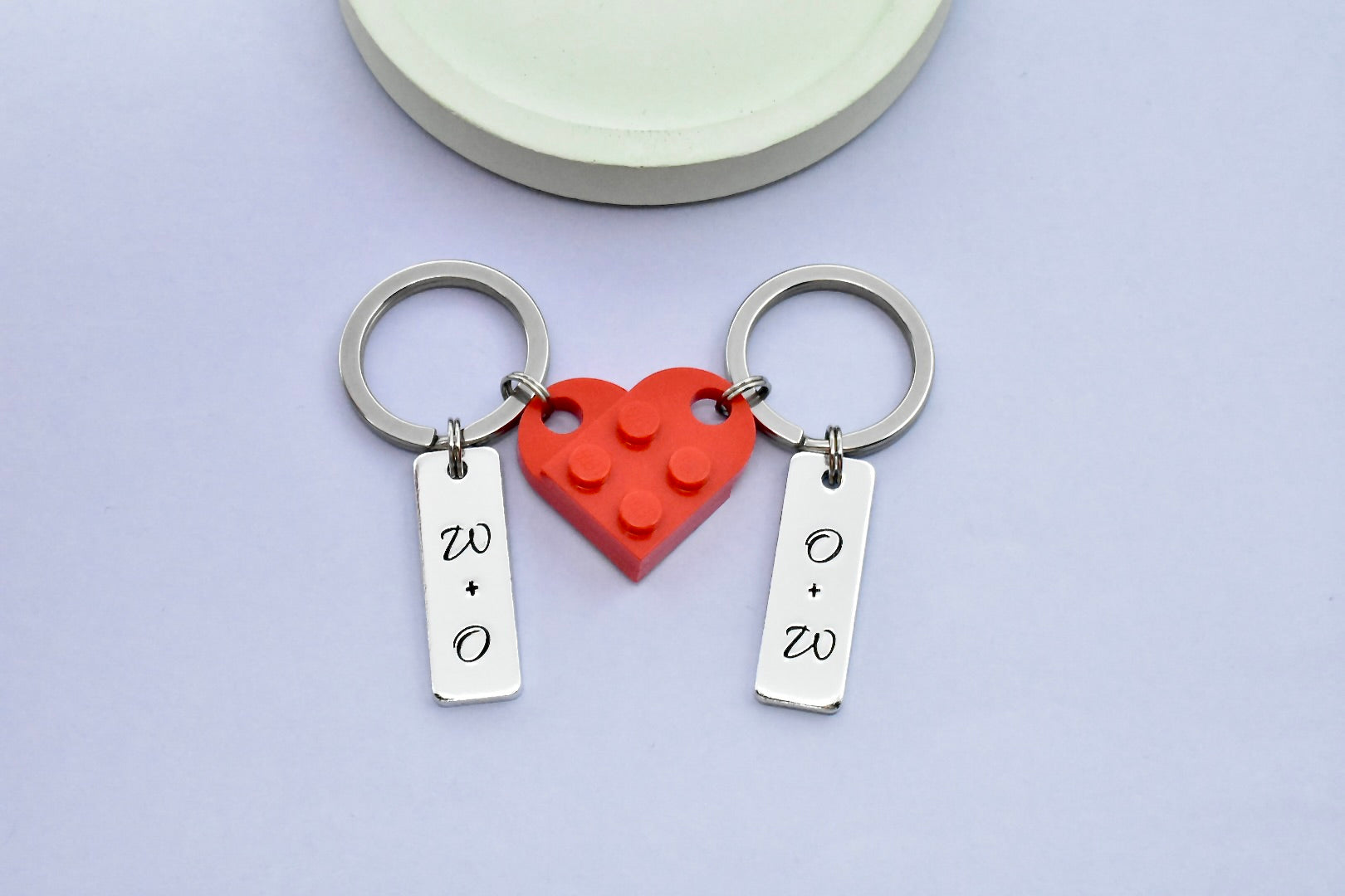 A set of heart-shaped connectable keyrings made of red plastic building blocks, each half of the heart has a small silver aluminium rectangle charm, personalised with couple's initials on each piece 