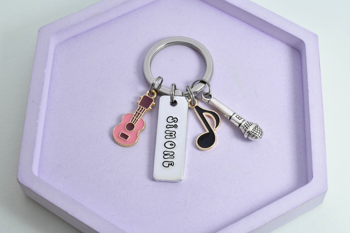 A personalised aluminium keyring with a rectangular tag, attached guitar, music note, and microphone charms, displayed on a purple surface.