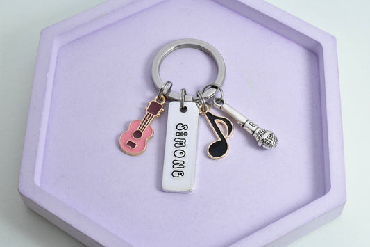A personalised aluminium keyring with a rectangular tag, attached guitar, music note, and microphone charms, displayed on a purple surface.