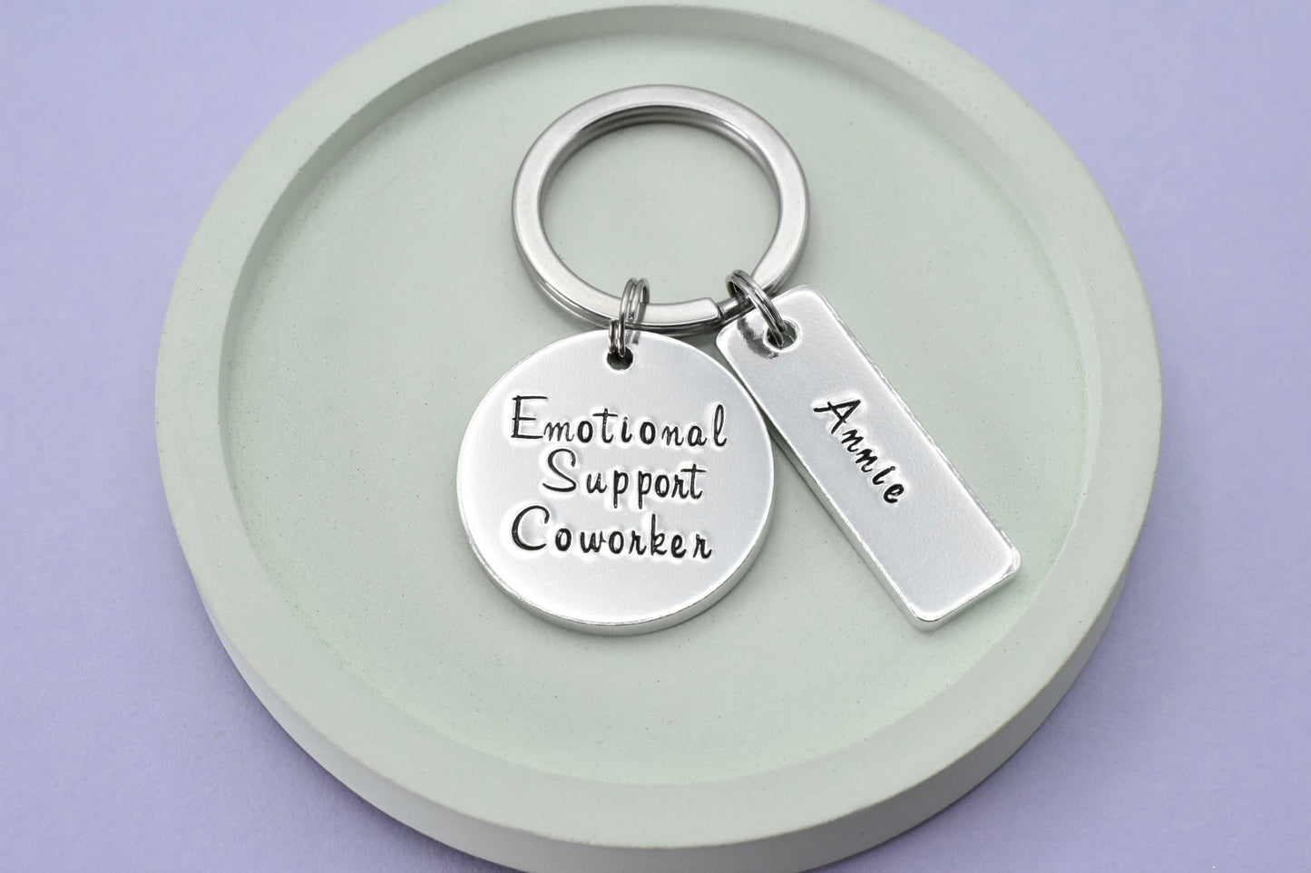 A keyring with a circular pendant and a small rectangular tag, with the text 'Emotional Support Coworker' and the name 'Annie'. The items are silver in color and the tag is attached to a keyring 