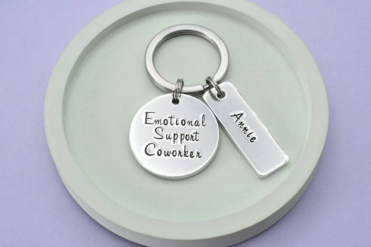 A keyring with a circular pendant and a small rectangular tag, with the text 'Emotional Support Coworker' and the name 'Annie'. The items are silver in color and the tag is attached to a keyring 