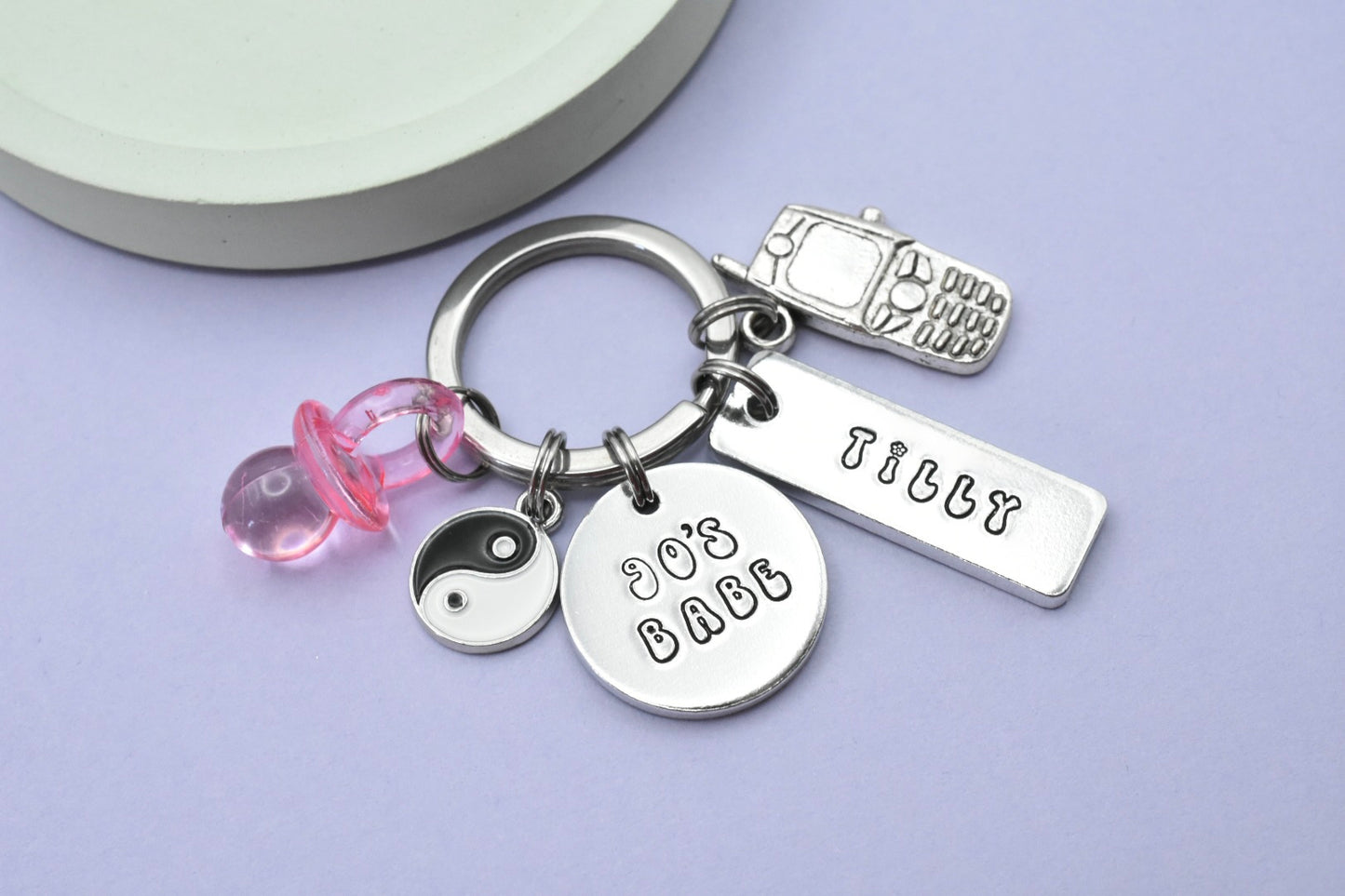 A keyring with multiple charms including a 90's dummy, a ying yang and a mobile phone charm. With a circular tag with the text '90's BABE'. The keyring and charms are silver, and the rectangular tag has a name stamped on it.
