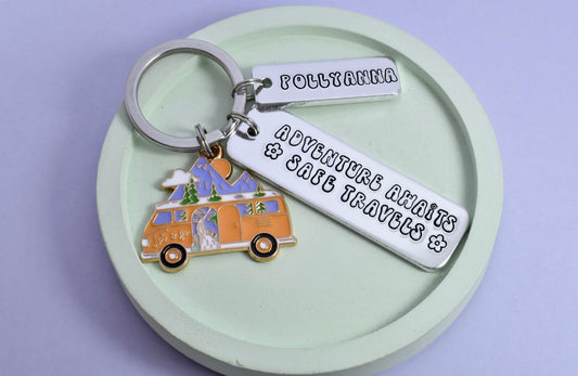 A personalised keyring with a rectangular tag that reads 'ADVENTURE AWAITS, SAFE TRAVELS", with personalised smaller rectangle and a charm shaped like a camper van. The keyring is attached to a silver-colored loop and is displayed against a green background.