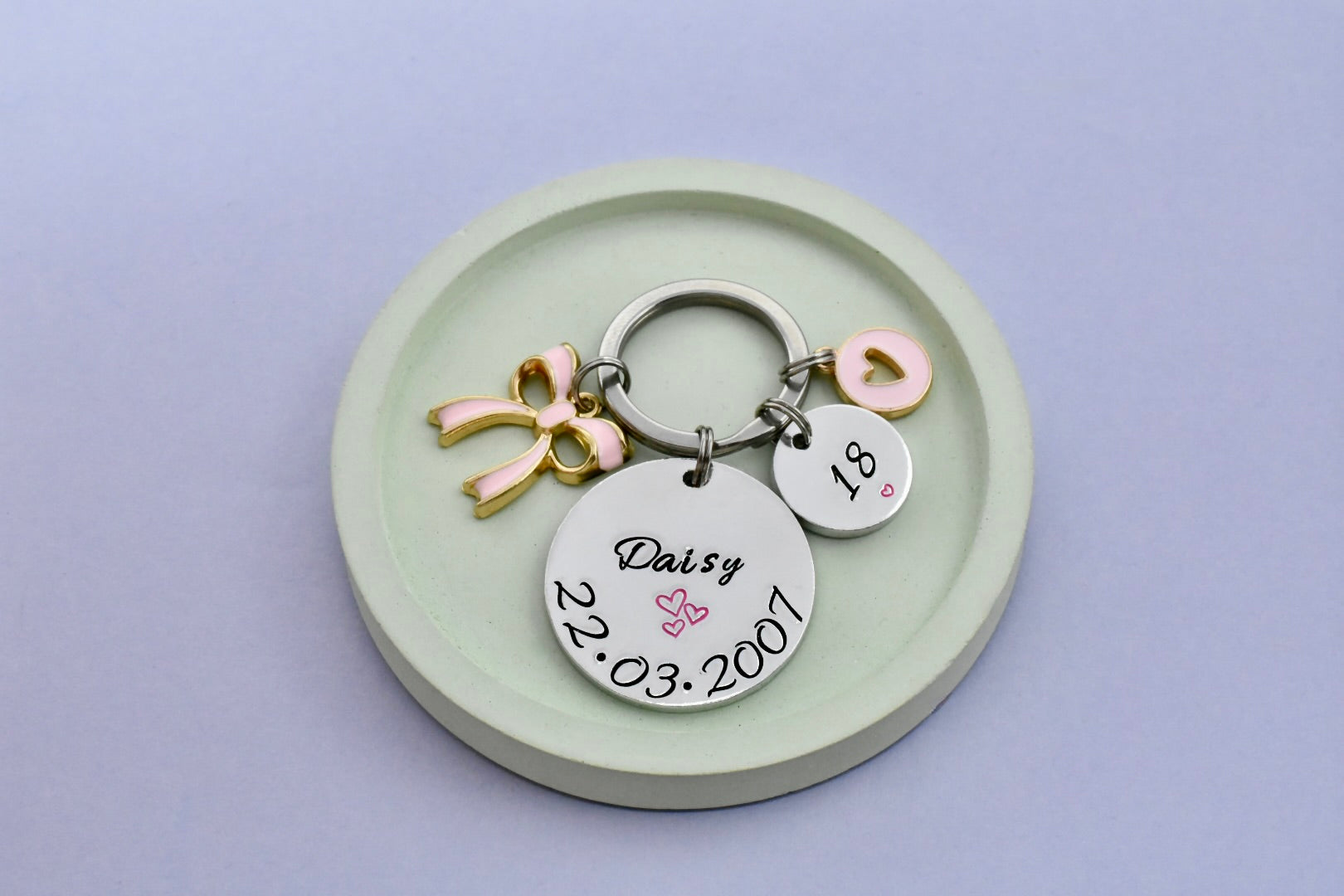 Personalised keychain with engraved birthday details with pink bow and heart charms on a light purple background