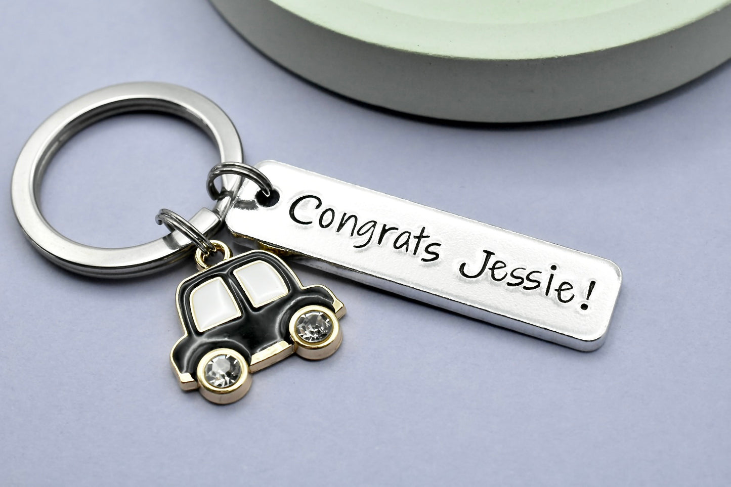 A silver-colored aluminium keyring with a personalised stamping that reads 'CONGRATS JESSIE!' alongside a small black colored car charm.