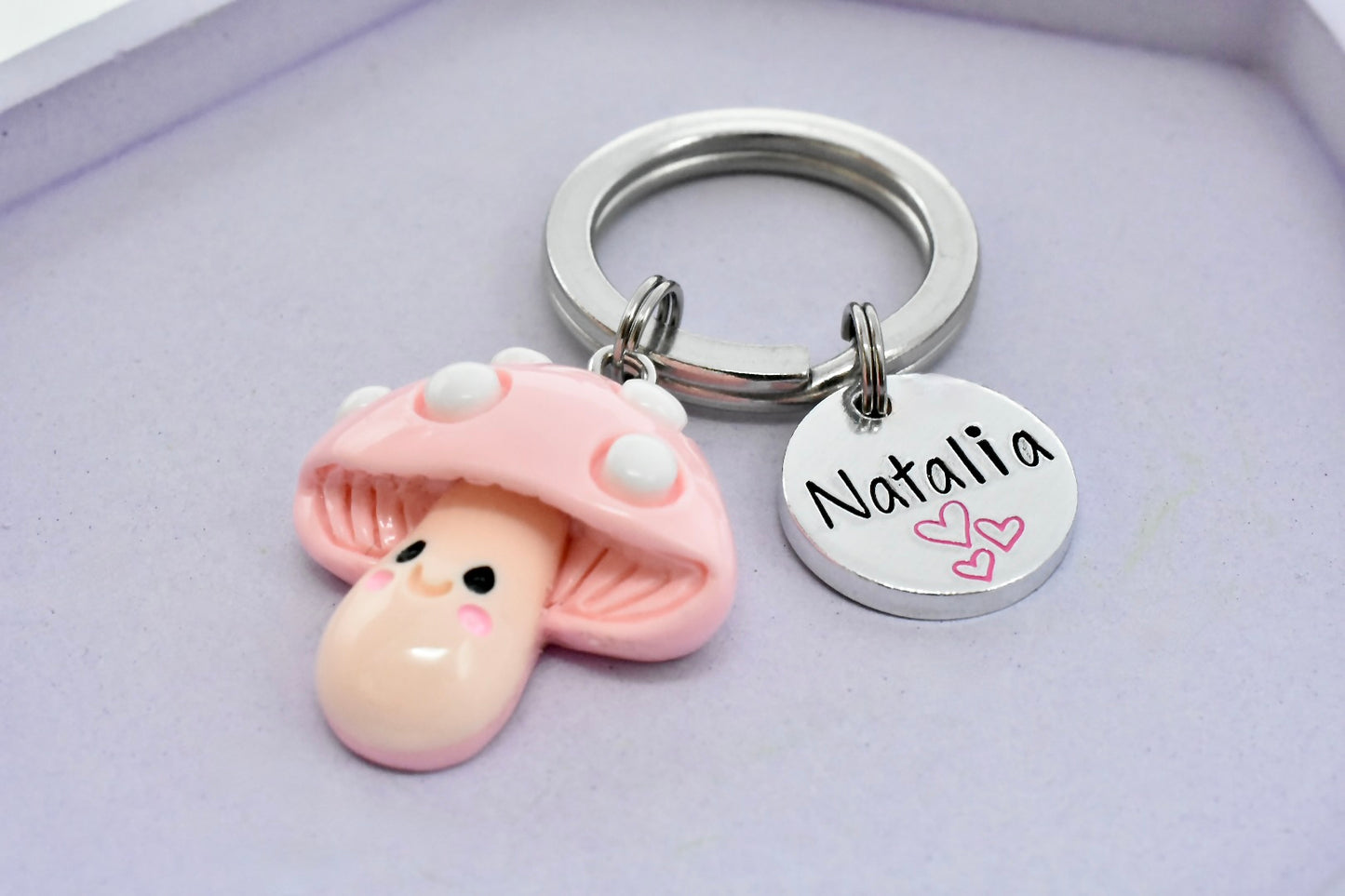 Keychain with a pink mushroom character charm and a round tag with 'Natalia' on a light purple background