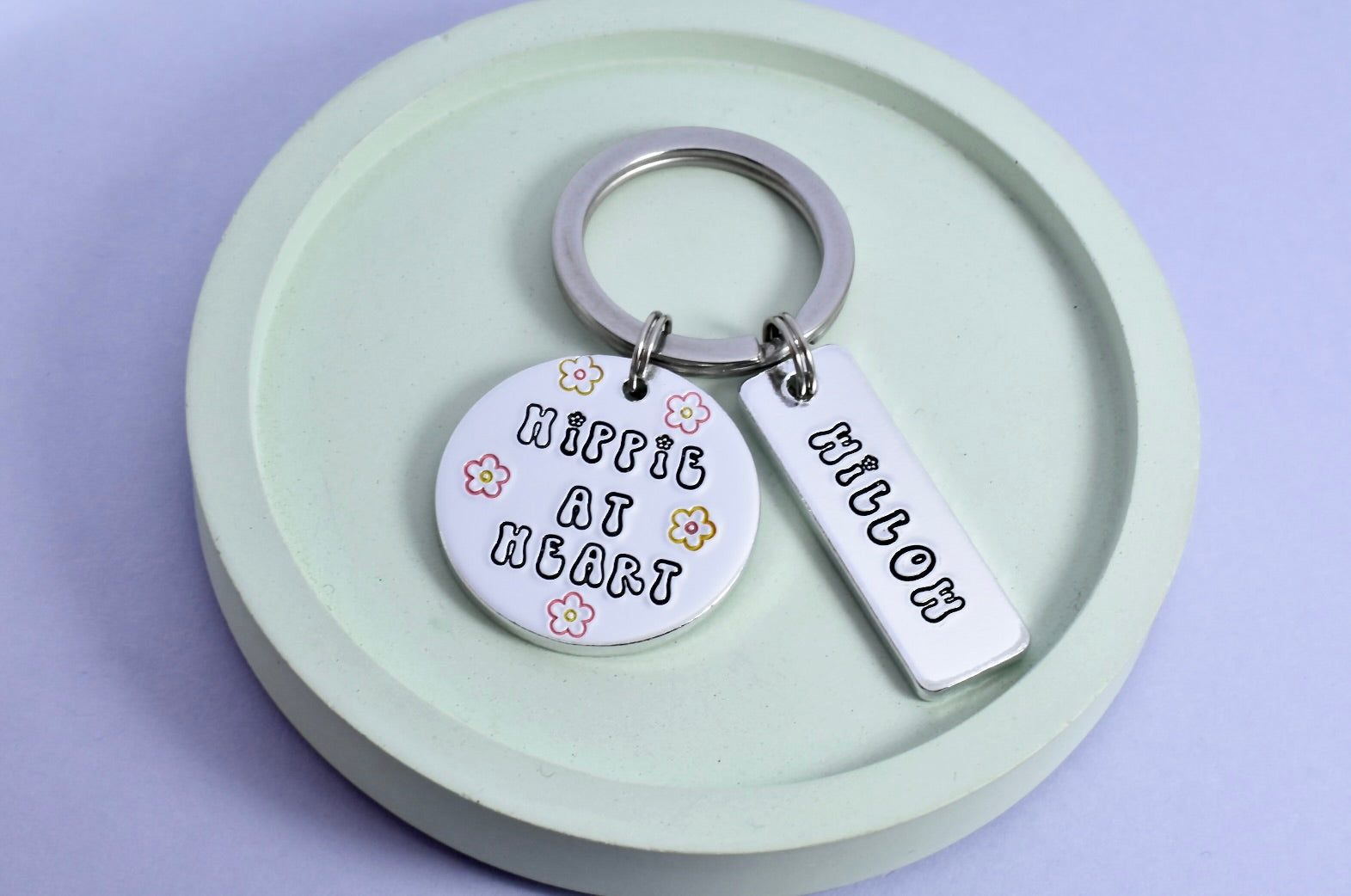 A personalized hippie-themed keyring with a floral design and the text 'Hippie At Heart' with small silver rectangle tag that is personalised with a name in bubble writing