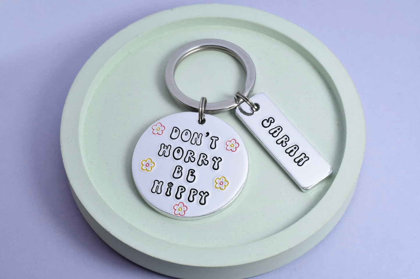 A silver aluminium keyring with a circular charm that has 'Don't Worry Be Hippie' printed on it, displayed on a green dish. There is also a smaller rectangular charm personalised with a  name