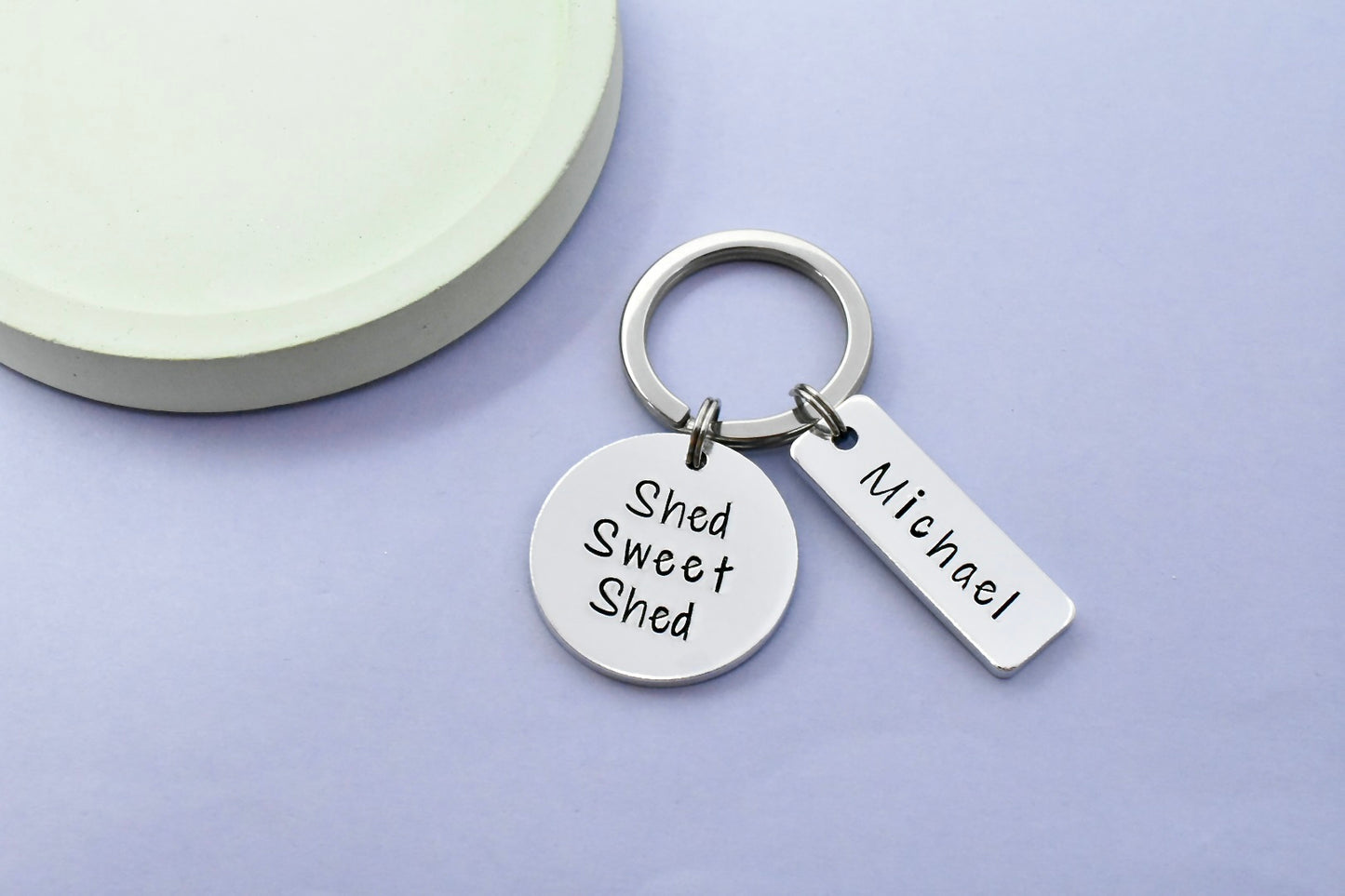 A keyring with a larger circular disc stamped with 'Shed Sweet Shed' and a smaller rectangular tag with the name 'Michael' attached to a split ring.
