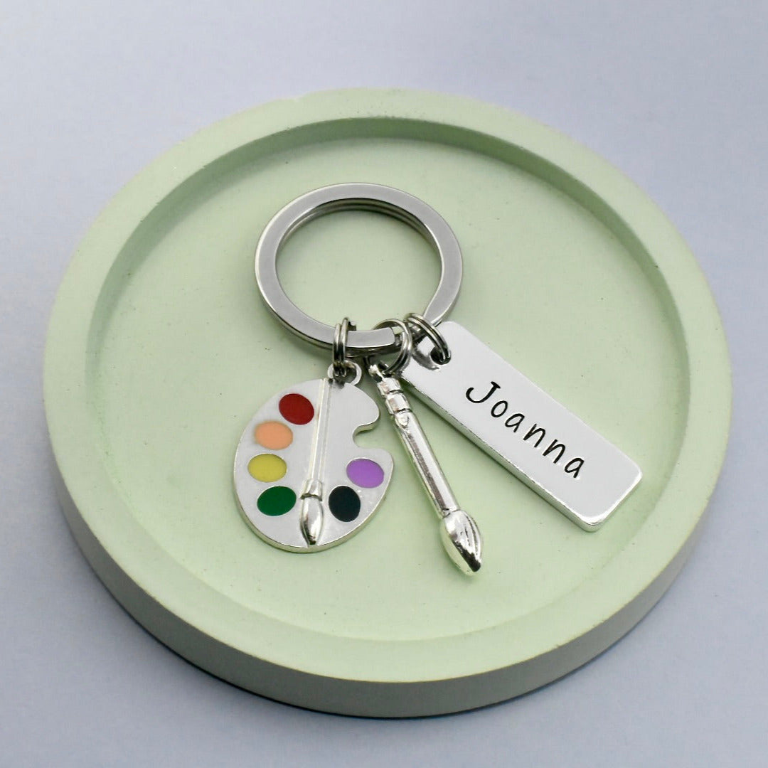 A personalised keyring with a painter theme, featuring a small aluminium rectangle, a paintbrush charm, and a paint pallet charm, all attached to a split ring. The keyring is hand stamped with the name 'Joanna' and presented on a round, green platform.