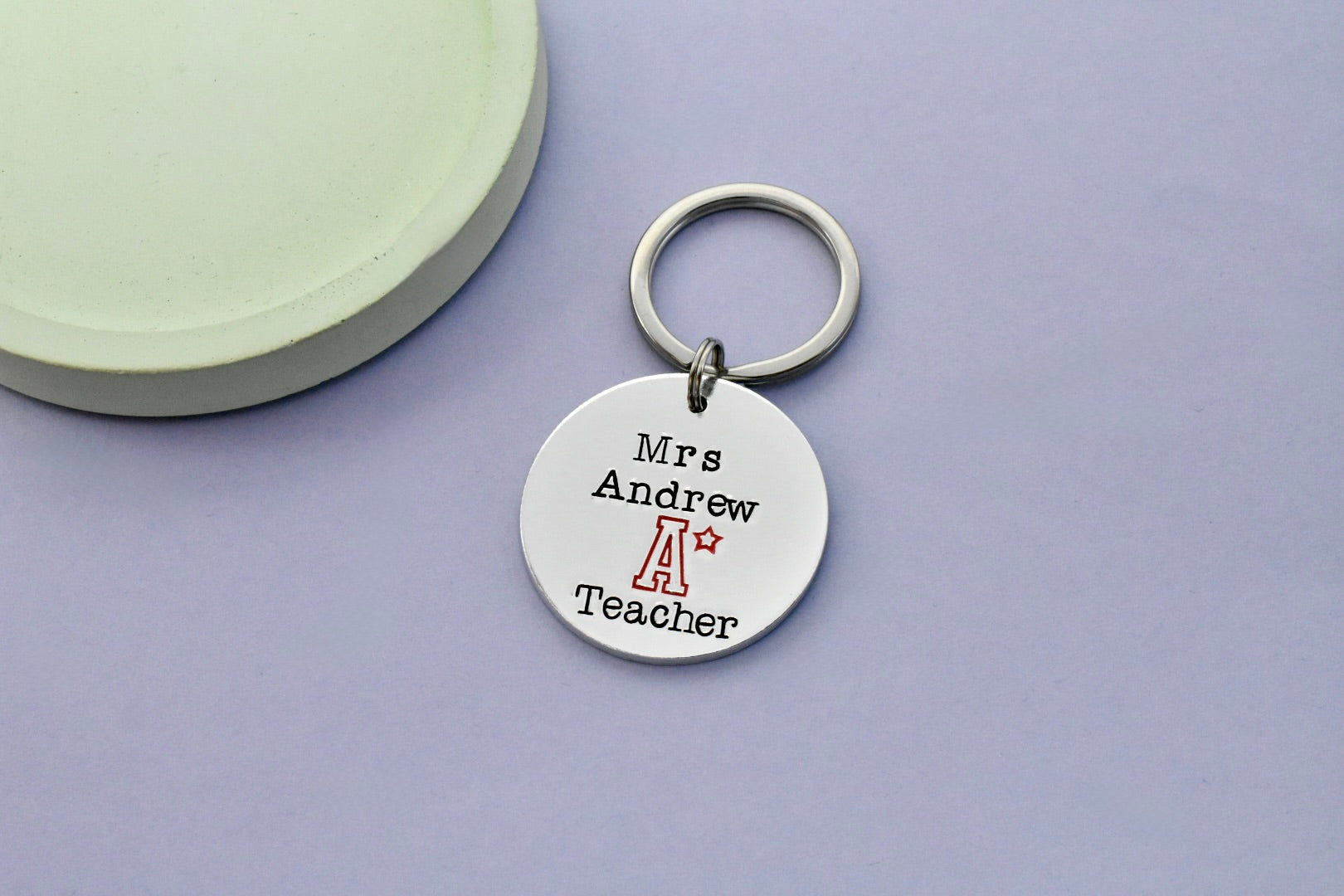 Aluminium keyring with 'Mrs. Andrew A Star (Stsr symbol) Teacher' hand stamped resting on a purple surface