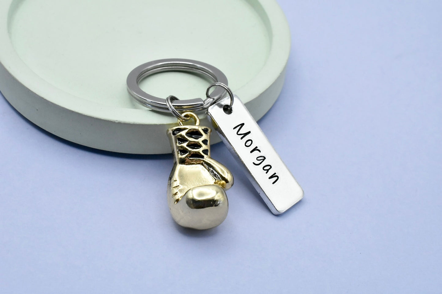 A personalised boxing keyring with a gold-colored 3D boxing glove charm and a rectangular tag with the name 'Morgan' engraved on it, presented on a purple background.