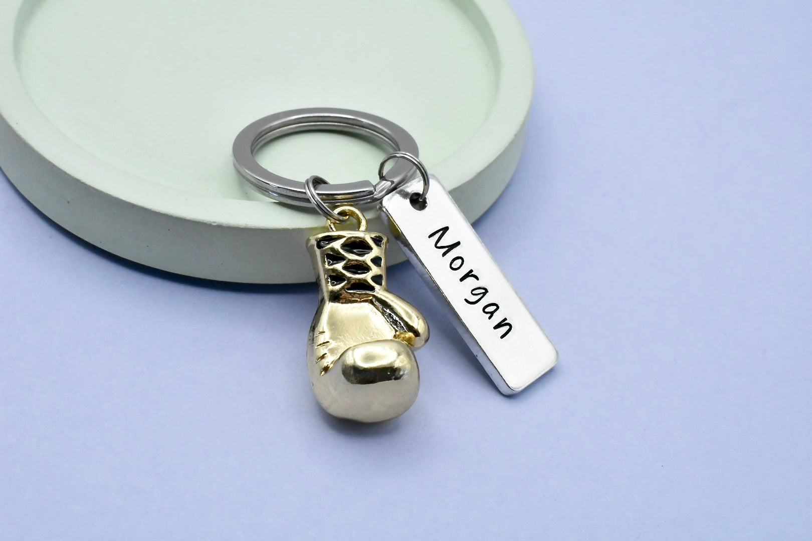A personalised boxing keyring with a gold-colored 3D boxing glove charm and a rectangular tag with the name 'Morgan' engraved on it, presented on a purple background.