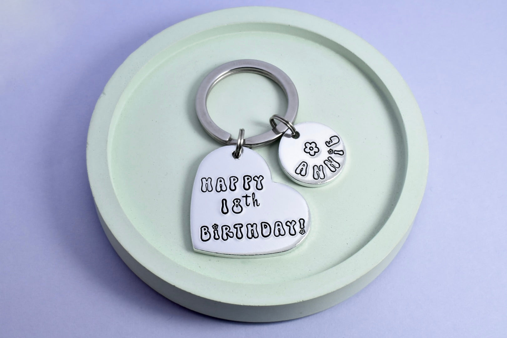 A silver heart shaped keyring with a smaller circular disc. Heart says "Happy 18th Birthday" in bubble writing. Small disc is personalised with a name and small black flower design