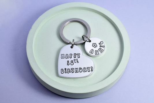 A silver heart shaped keyring with a smaller circular disc. Heart says "Happy 18th Birthday" in bubble writing. Small disc is personalised with a name and small black flower design