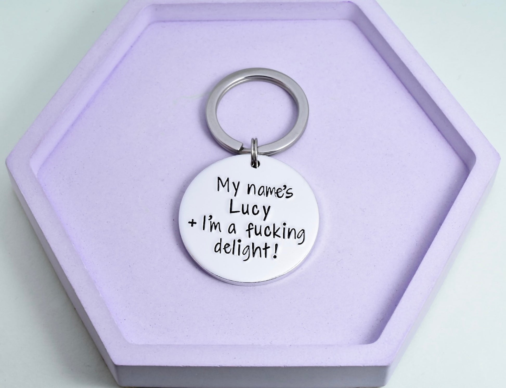 A personalized keyring with a split ring, engraved with the text 'MY NAME'S (CUSTOMISABLE) + I'M A FUCKING DELIGHT!' on a purple background, placed on a purple hexagonal box.