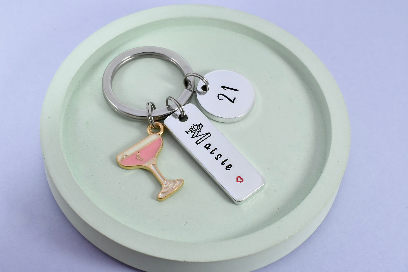 A personalised birthday keyring with a champagne cocktail charm, customised with a name and age on a rectangular tag, presented on a light green aluminium keyring.