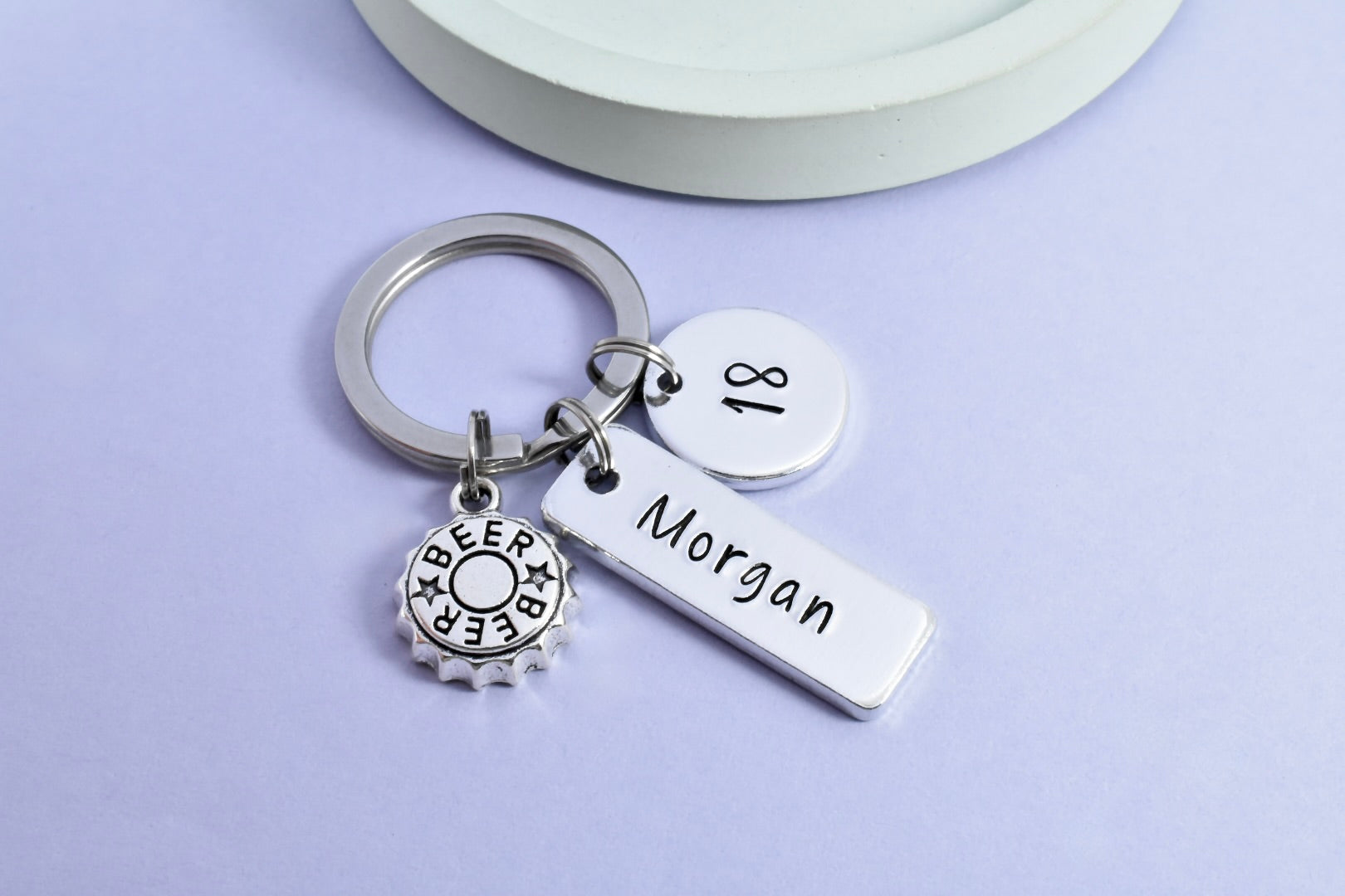 A personalised aluminium keyring with a beer cap charm, customised with the name 'Morgan' and the age '18'. It includes a rectangular tag and a disc charm with the name and age, attached to a split ring.