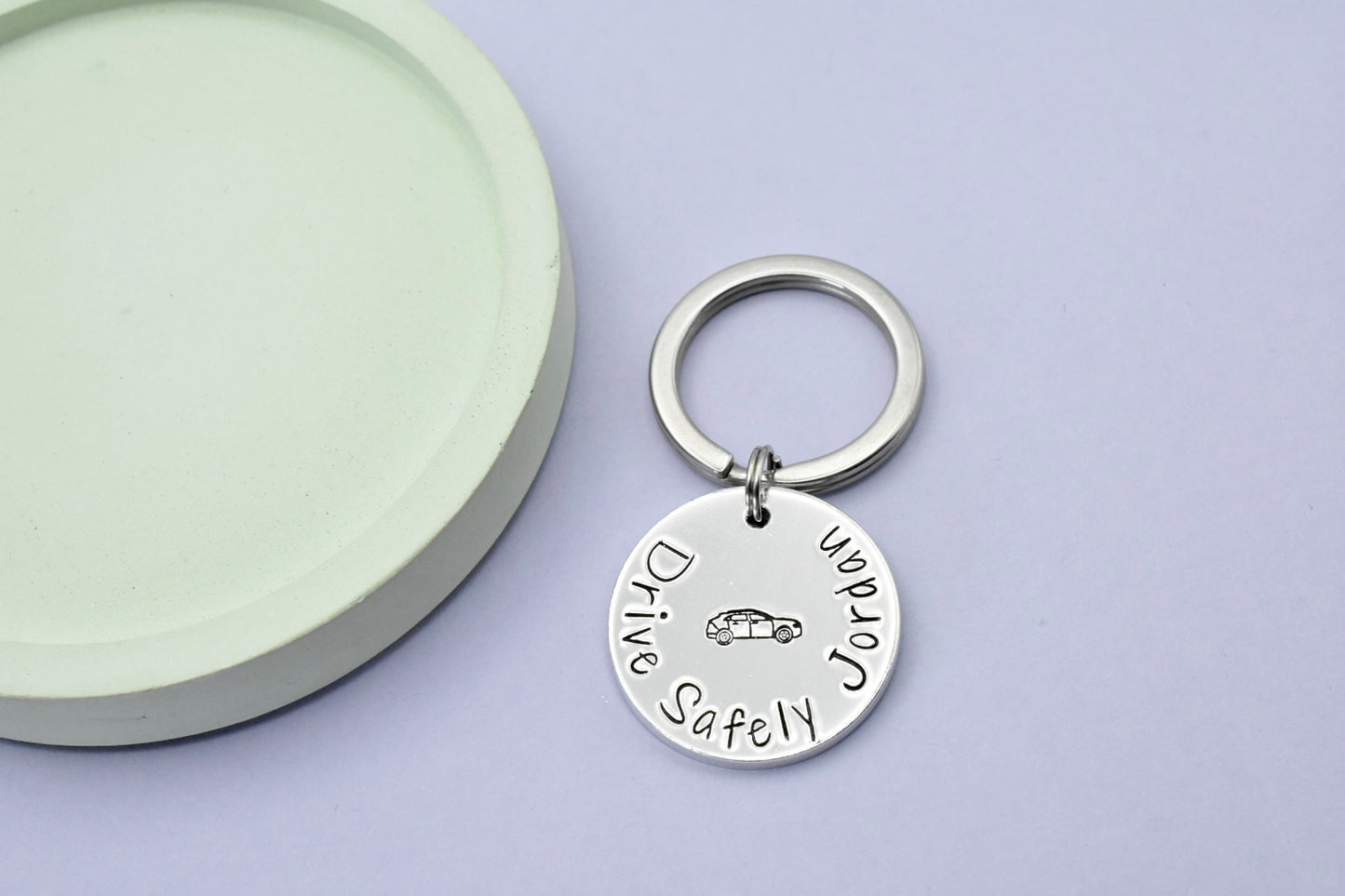 A silver-colored keyring with a disc-shaped charm personalised with 'Drive Safely Jordan' on it and car design in the center, attached to a split ring. The keyring is presented against a light background.