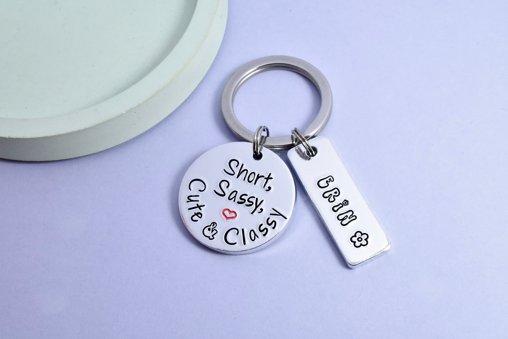 A silver aluminium keyring with a circular charm featuring the text 'Short, Sassy, Cute & Classy', and a smaller rectangular charm personalised with tand name and a flower design. The keyring is displayed against a purple background.