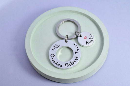 A personalised keyring with 'This Grandma Belongs To' inscribed on a aluminium washer, attached to a split ring, includes a small silver disc with a heart symbol, personalised with a grandchild's name