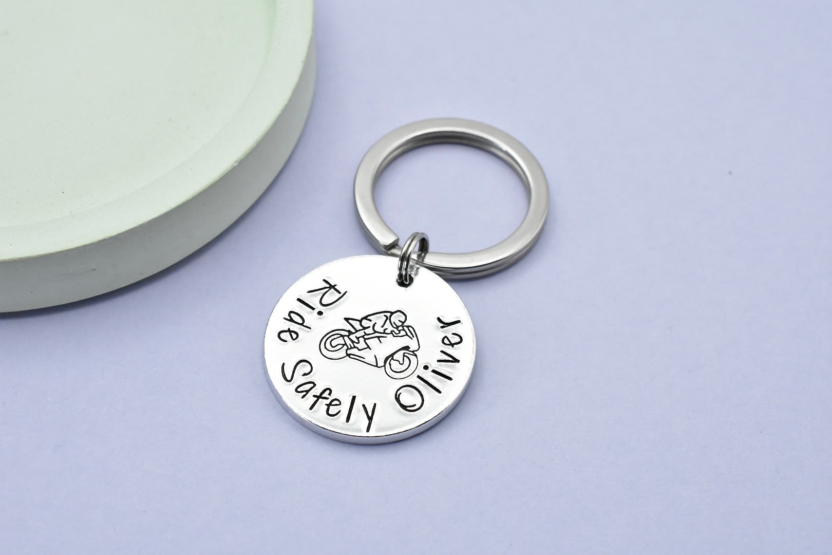 A personalised keyring with a circular aluminium disc, featuring text "Ride Safely Oliver" and a motorcycle design, attached to a split ring, displayed against a neutral background.