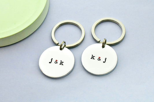 Pair of keyrings, made of aluminium, has initials on t that are separated by a heart ampersand