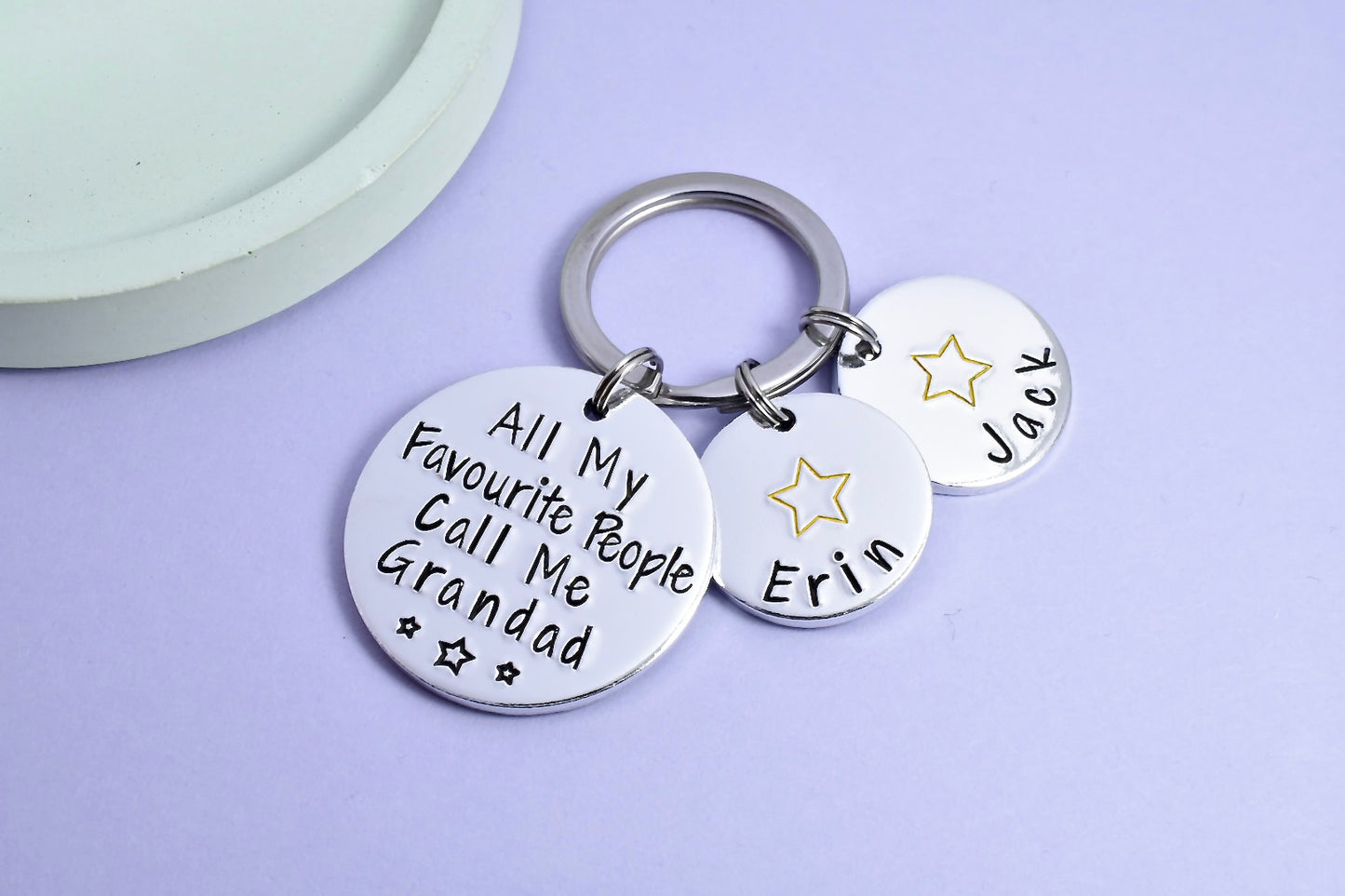 A custom aluminium keyring with the phrase 'All My Favourite People Call Me Grandad' followed by personalised names 'Erin' and 'Jack' on smaller discs, against a purple background.