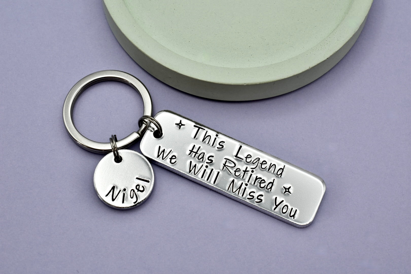 A personalised retirement keyring with a large rectangular aluminium tag stamped with 'This Legend Has Retired. We Will Miss You' and a smaller disc personalised with a name.