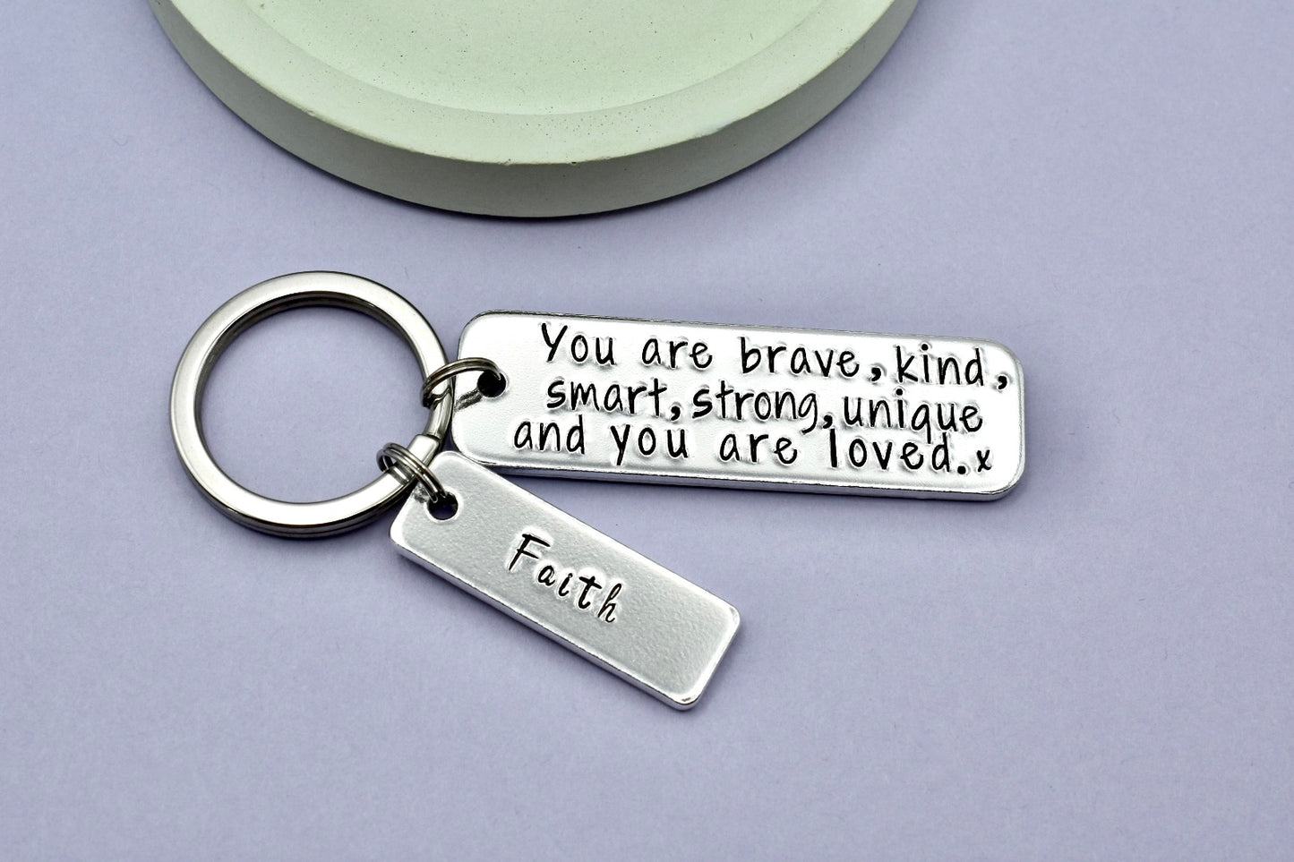 A silver keyring with two rectangular tags, one with the personalized name 'Faith' and the other with an inspirational message 'You are brave, kind, smart, strong, unique and you're loved.'.