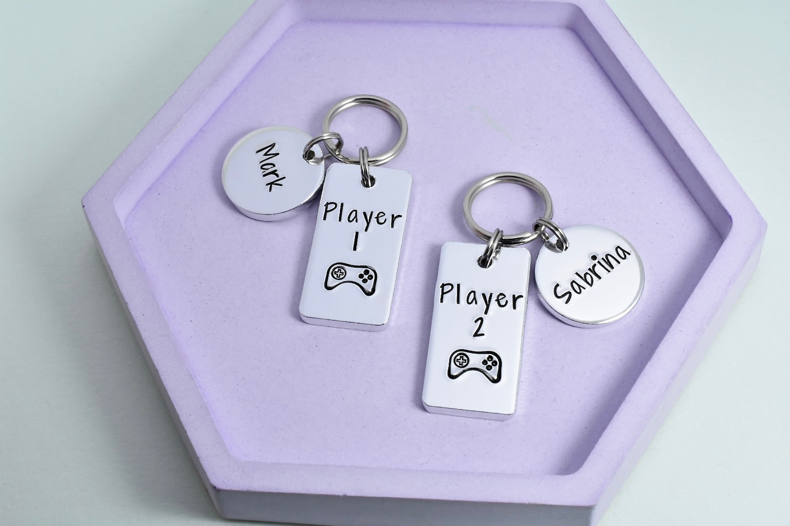Two personalised aluminum keyrings with 'PLAYER 1' and 'PLAYER 2' text, along with a small round name disc, all on a purple background. 