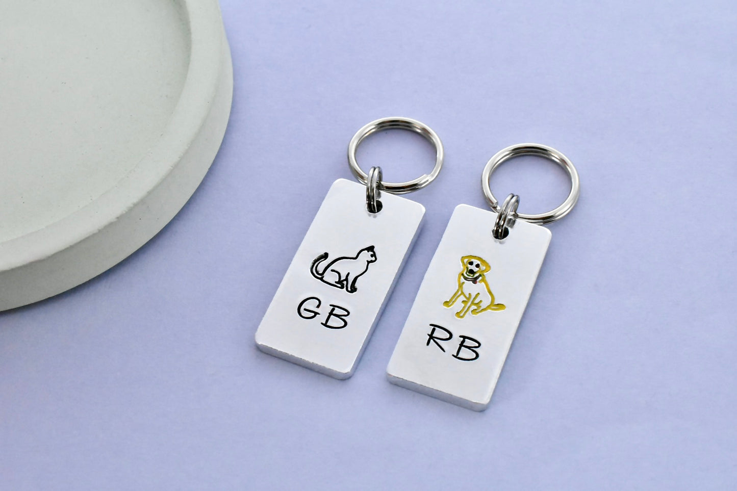 Two small rectangle silver aluminium keychains with hand stamped designs, one a black cat and the other a yellow labrador, with couple's initials on each piece