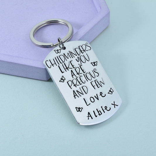 A personalised metal keyring, hand stamped with "Childminders like you are precious and few" and a child's name, with butterfly designs. displayed on a purple surface. 
