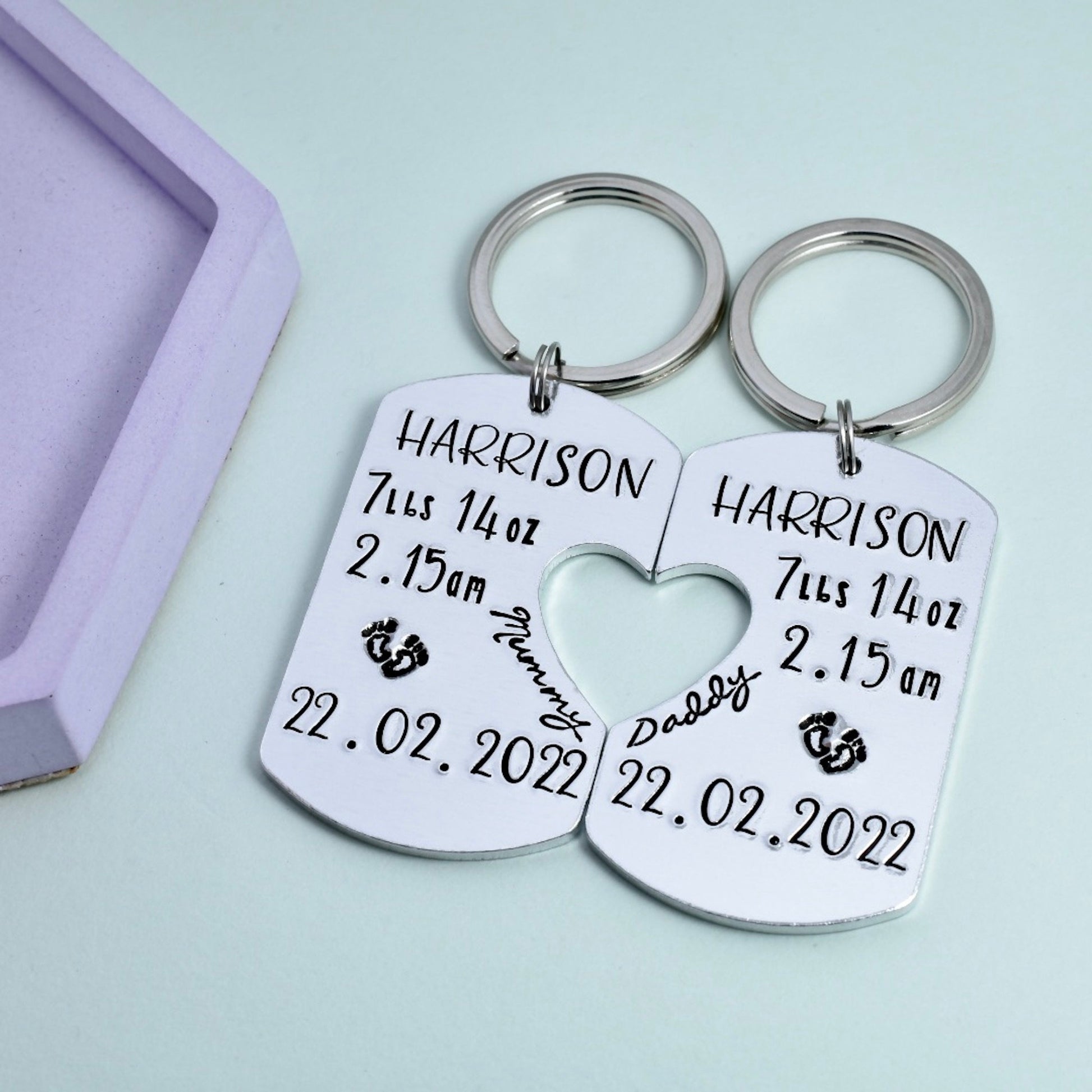 Two aluminium keyrings attached to a keychain, with personalised stampings including a name and birth details. One keyring is engraved with 'Mummy' and the other with 'Daddy', both dated and with heart cut-outs in the center. Keyring set for a new Mummy and Daddy