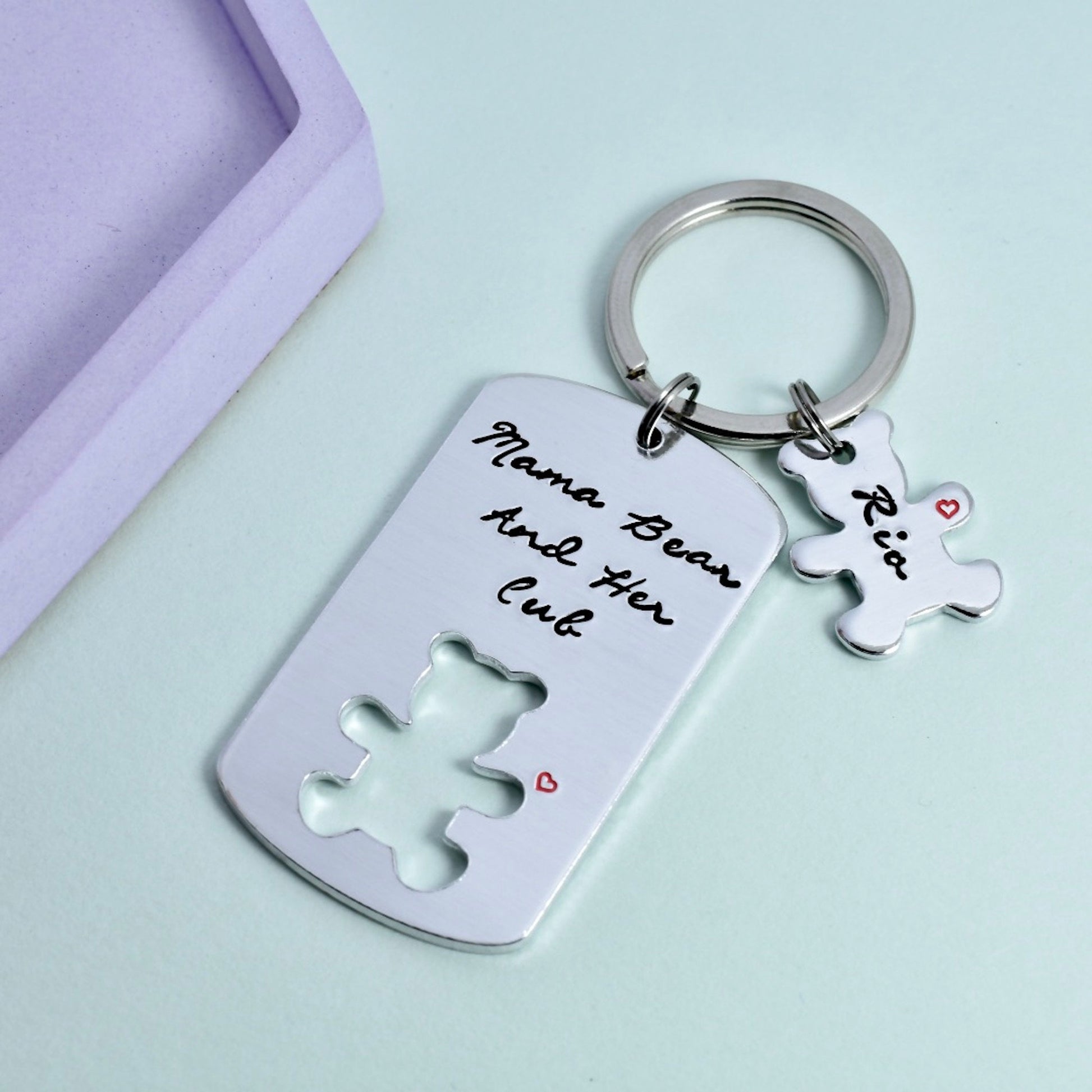 A personalised keyring with a bear and her cub design, with the inscription 'MAMA BEAR AND HER CUB' on a silver-colored ring. There is a small bear-shaped name charm attached to the ring.