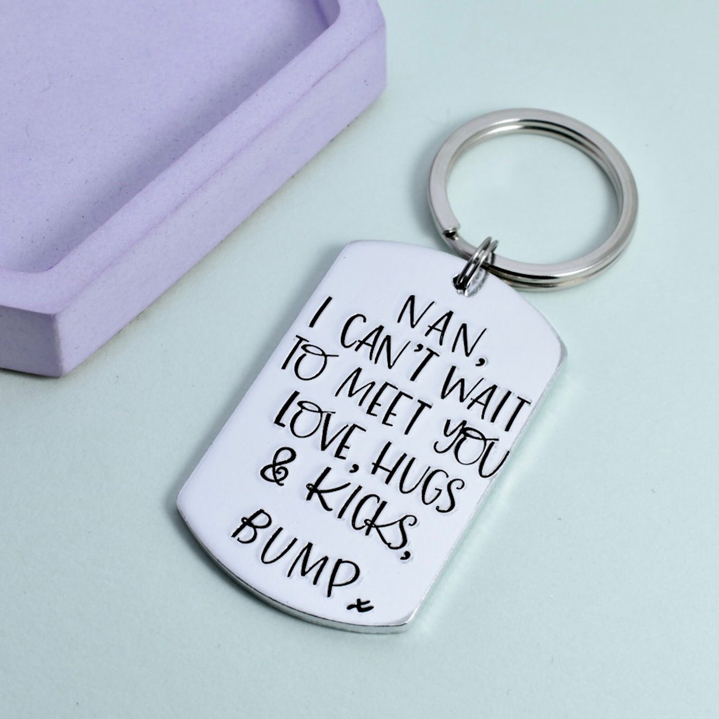 A personalized aluminum keyring with the text 'I CAN'T WAIT TO MEET YOU LOVE, HUGS & KICKS BUMP' engraved on it, alongside a small 'x' symbol, presented on a light background,  indicating a gift from an unborn child to a parent.