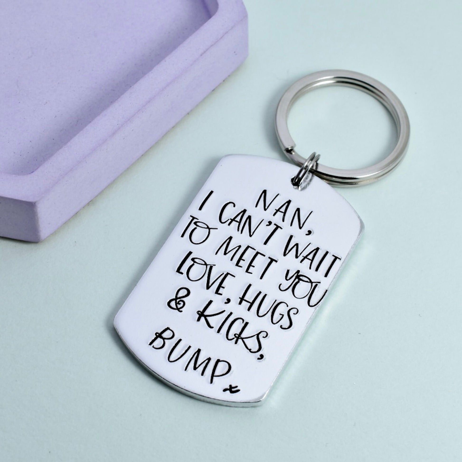 A personalized aluminum keyring with the text 'I CAN'T WAIT TO MEET YOU LOVE, HUGS & KICKS BUMP' engraved on it, alongside a small 'x' symbol, presented on a light background,  indicating a gift from an unborn child to a parent.