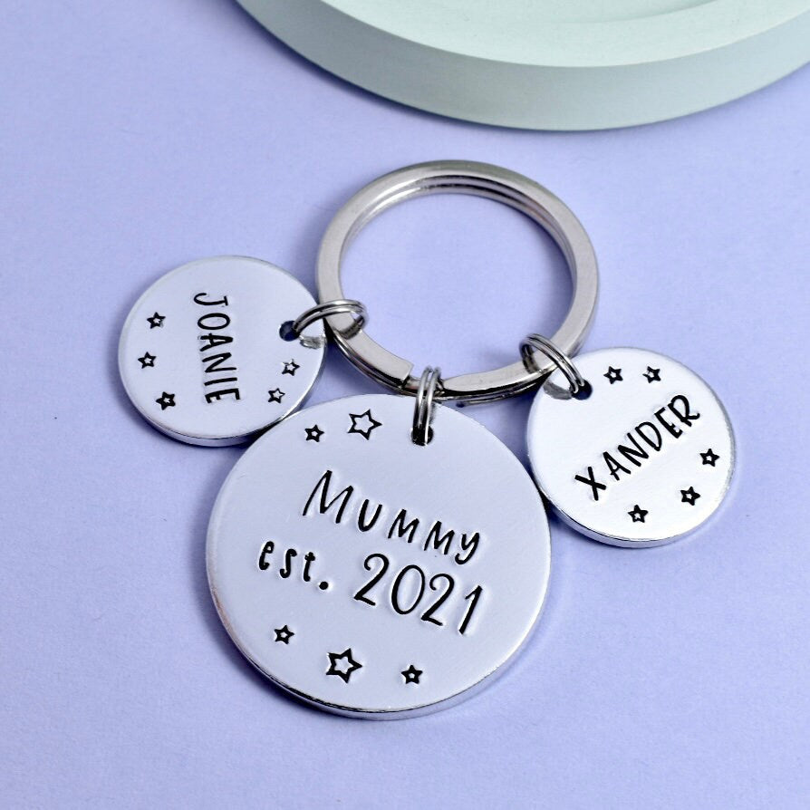 A personalised keyring gift set with three round aluminium discs, one large and two small, attached to a keychain. The large disc reads 'Mummy est.