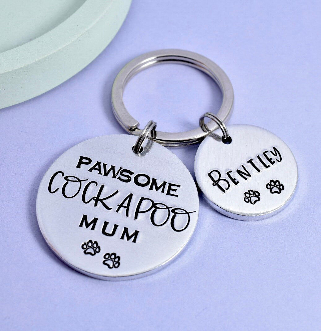 A personalised keyring with two aluminum discs, one larger and one smaller, with the text 'PAWSOME COCKAPOO MUM' and a smaller engraved dog name 'Bentley'.