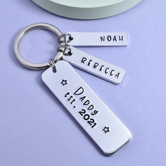 A personalised keyring with three silver aluminium rectangles, one large and two small, personalised with the names 'NOAH' and 'REBECCA'. The large rectangle has 'Daddy est.