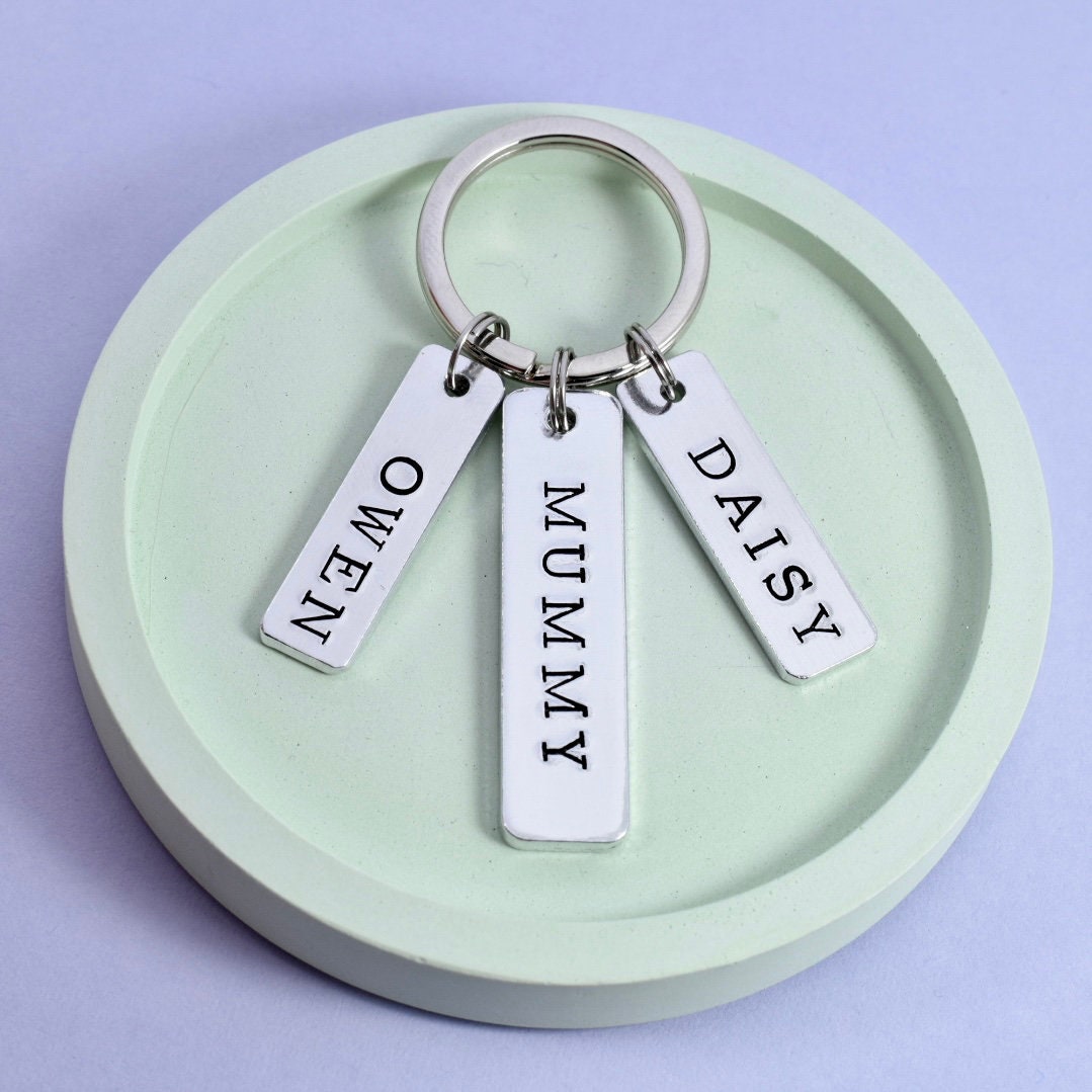 A personalised keyring with three aluminum tags, one larger and two smaller, each with a child's name written on it. The larger tag has 'MUMMY' written on it, 