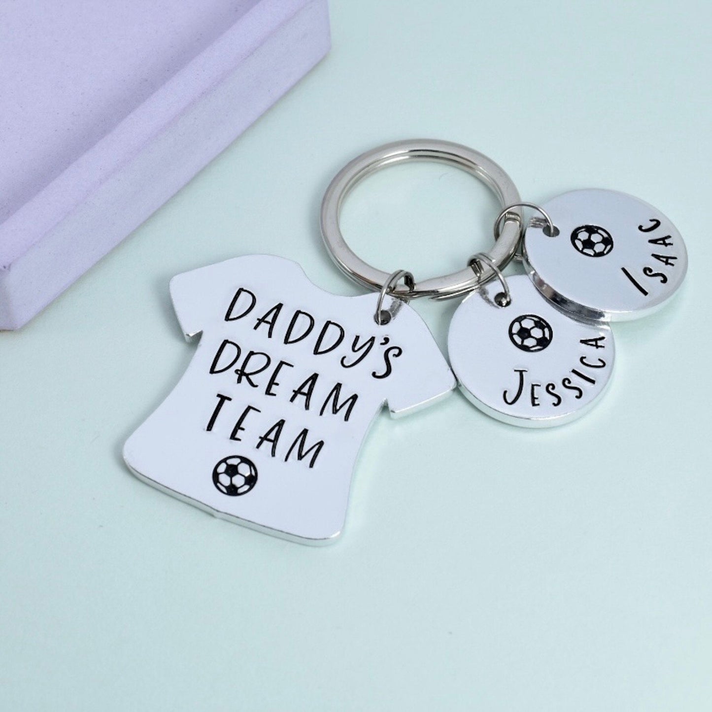 A personalised football dad keyring with a shirt-shaped keychain engraved with 'DADDY'S DREAM TEAM' and circular name tags attached to a split ring, displayed on a purple background.