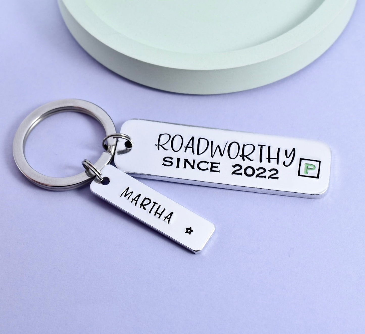 A personalised keyring with a larger rectangle stamped with 'ROADWORTHY SINCE 2023' and a smaller rectangle personalised with a name. Both have a mock green P plate design. New driver name is stamped on smaller rectangle charm
