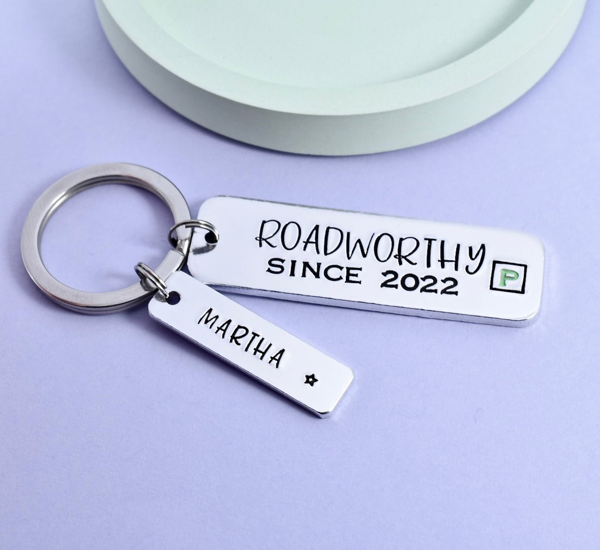 A personalised keyring with a larger rectangle stamped with 'ROADWORTHY SINCE 2023' and a smaller rectangle personalised with a name. Both have a mock green P plate design. New driver name is stamped on smaller rectangle charm