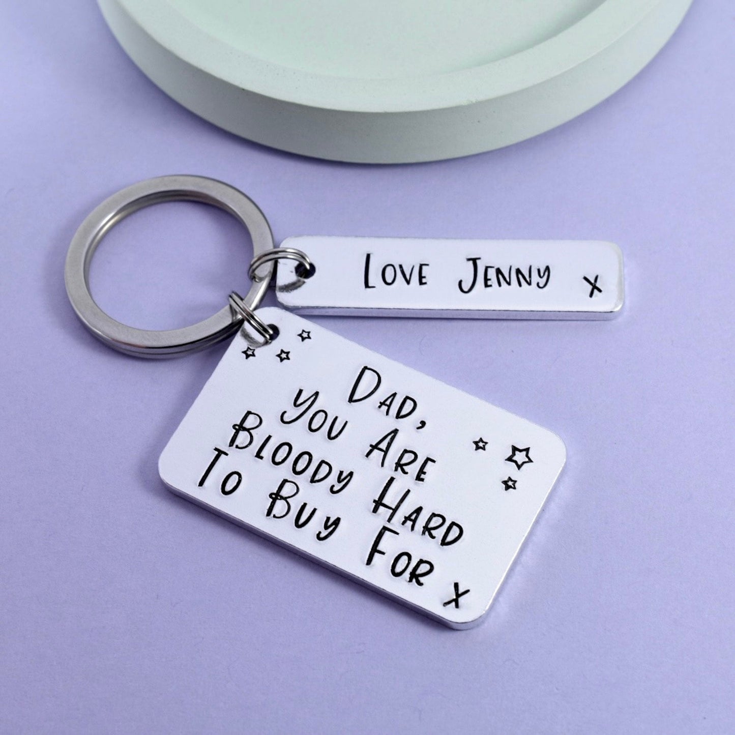 A silver aluminium keyring with two rectangular tags, one larger than the other. The larger tag has a humorous message stamped on it, "DAD YOU ARE BLOODY HARD TO BUY FOR" and the smaller tag is personalised with "love Jenny"