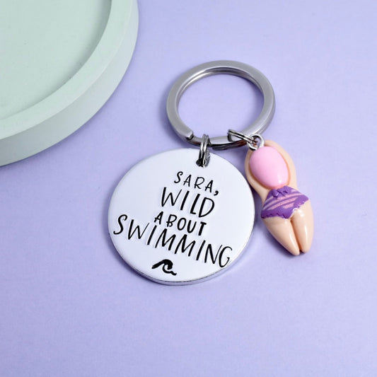 A personalised aluminium keyring with the phrase 'SARA'S WILD ABOUT SWIMMING' written on it, accompanied by a small wave detail and a charm in the shape of a swimmer wearing a purple swimsuit. cold water swimmer