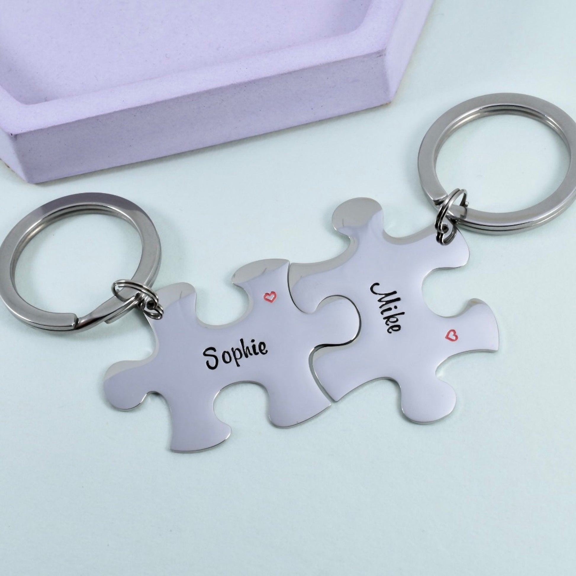 A pair of interlocking steel puzzle piece keyrings with names 'Sophie' and 'Mike' stamped on them, alongside a small heart symbol. The keyrings are silver-colored and have a shiny finish and are personalised
